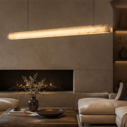 A modern living room interior with a long horizontal linear pendant light glowing above a fireplace. The room features textured concrete walls, a beige sectional sofa, a wooden coffee table with a dark vase of branches, and built-in shelving with warm ambient lighting.