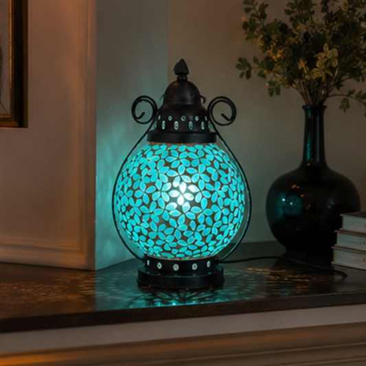A decorative table lamp with a glowing turquoise mosaic globe and black metal frame, placed on a mantel in a home interior next to a dark glass vase with flowers.