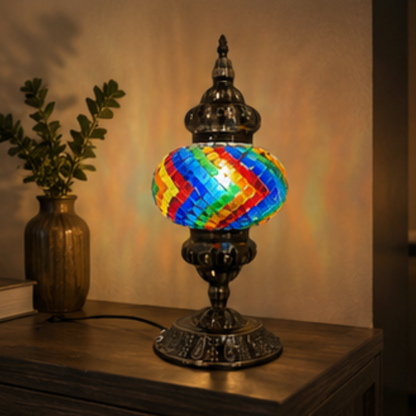 A decorative table lamp with an ornate dark metal base and a colorful mosaic globe shade featuring red, orange, yellow, green, and blue tiles, shown lit on a wooden console table in a warm indoor setting.