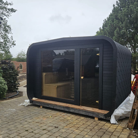 A modern black outdoor office or cabin with large glass doors and windows, situated on a paved driveway in a backyard setting.