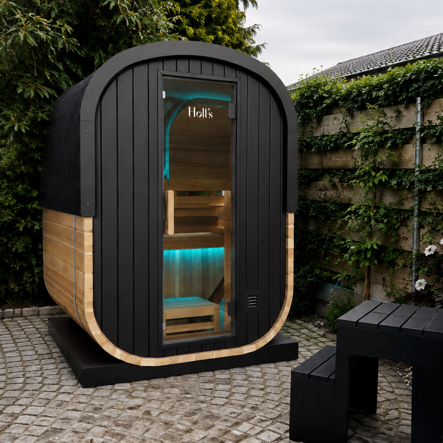 A modern outdoor sauna cabin with a black wood-look exterior, curved light wood trim, and a glass front door showing warm interior lighting, situated on a paved patio next to a hot tub in a garden setting.