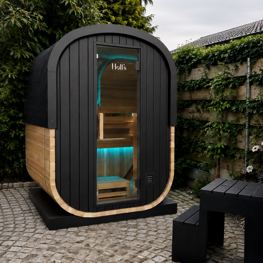 A modern outdoor sauna cabin with a black wood-look exterior, curved light wood trim, and a glass front door showing warm interior lighting, situated on a paved patio next to a hot tub in a garden setting.