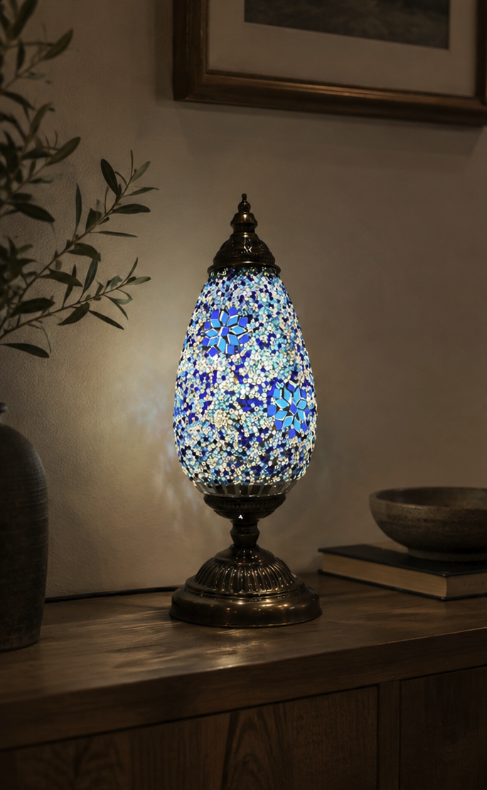Serena mosaic table lamp lit on a wooden console table, showing the blue and white stained glass shade and bronze base