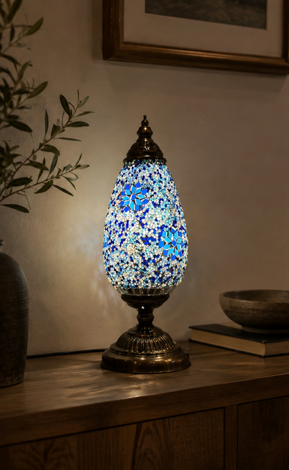 Serena mosaic table lamp lit on a wooden console table, showing the blue and white stained glass shade and bronze base