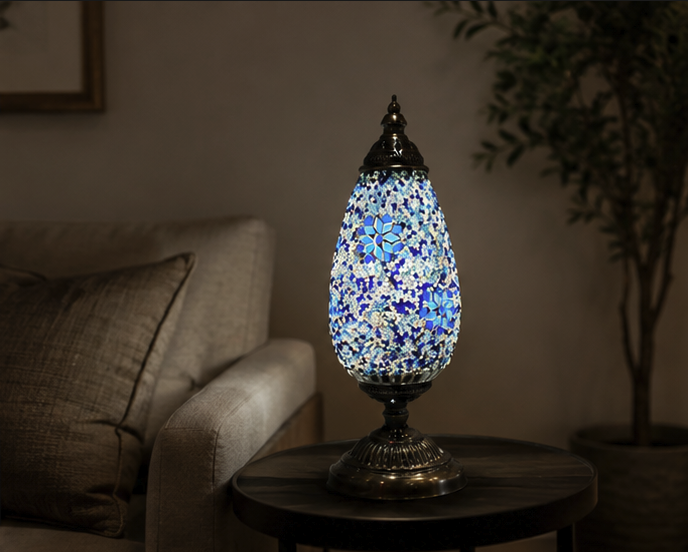 Serena mosaic table lamp glowing with blue, white and turquoise glass on a side table beside a sofa in a dimly lit living room